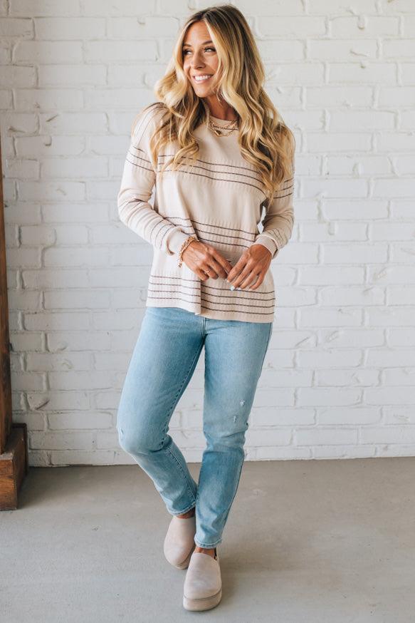 Woman wearing a striped sweater and light blue jeans against a white brick wall.
