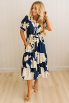 Woman wearing a navy and white floral dress against a light wooden paneled wall.