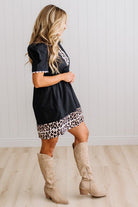 BLONDE WOMAN IN A BRIGHTLY LIT STUDIO WITH LIGHT WOOD FLOORS WEARING LIGHT TAN COWBOY BOOTS AND A BLACK KNEE LENGTH DRESS WITH LEOPARD PRINT ACCENT AND RICKRACK DETAILING.