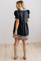 BLONDE WOMAN IN A BRIGHTLY LIT STUDIO WITH LIGHT WOOD FLOORS WEARING LIGHT TAN COWBOY BOOTS AND A BLACK KNEE LENGTH DRESS WITH LEOPARD PRINT ACCENT AND RICKRACK DETAILING.