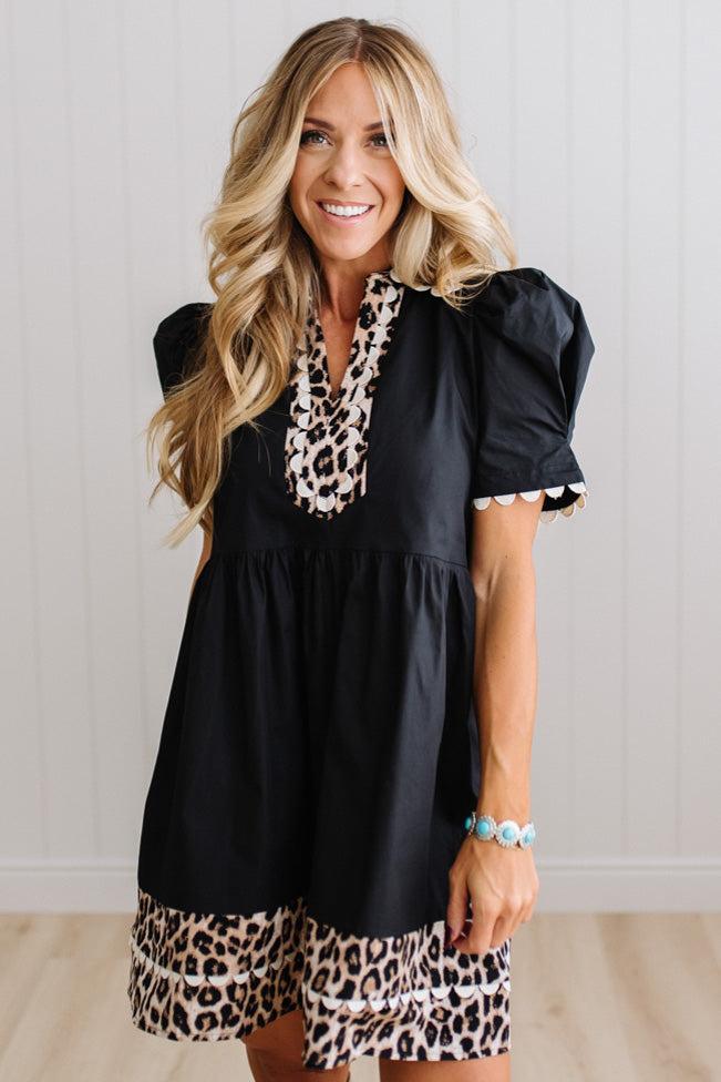 BLONDE WOMAN IN A BRIGHTLY LIT STUDIO WITH LIGHT WOOD FLOORS WEARING LIGHT TAN COWBOY BOOTS AND A BLACK KNEE LENGTH DRESS WITH LEOPARD PRINT ACCENT AND RICKRACK DETAILING.