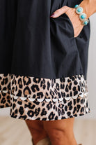 BLONDE WOMAN IN A BRIGHTLY LIT STUDIO WITH LIGHT WOOD FLOORS WEARING LIGHT TAN COWBOY BOOTS AND A BLACK KNEE LENGTH DRESS WITH LEOPARD PRINT ACCENT AND RICKRACK DETAILING.