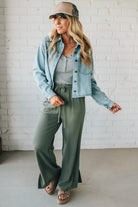 Woman wearing a denim jacket, gray top, and green pants against a white brick wall.