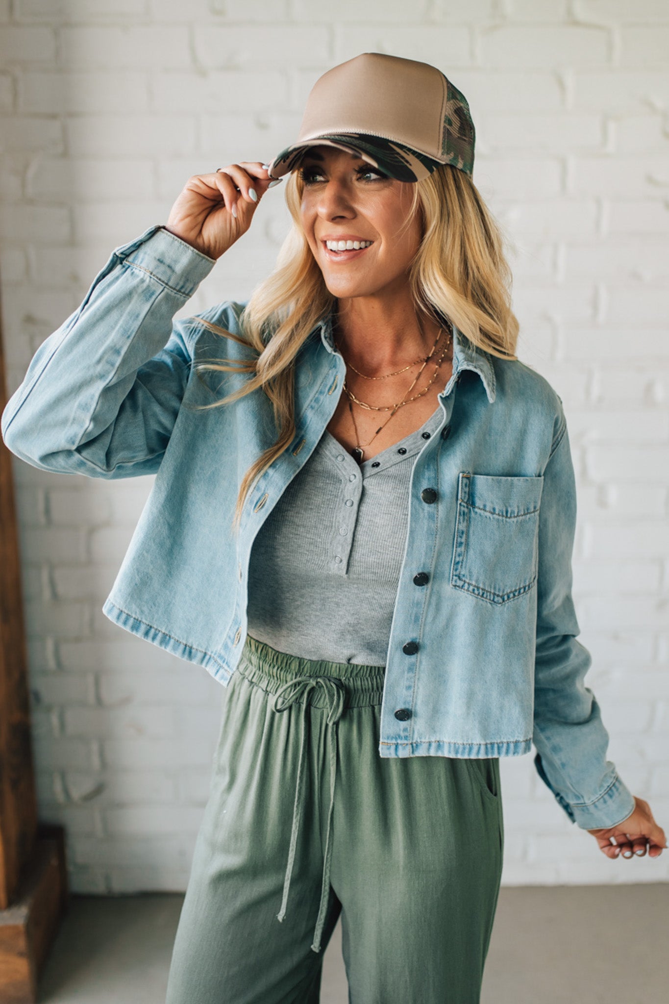 Woman wearing a denim jacket, gray top, and green pants with a beige cap indoors.