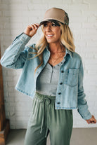 Woman wearing a denim jacket, gray top, and green pants with a beige cap indoors.