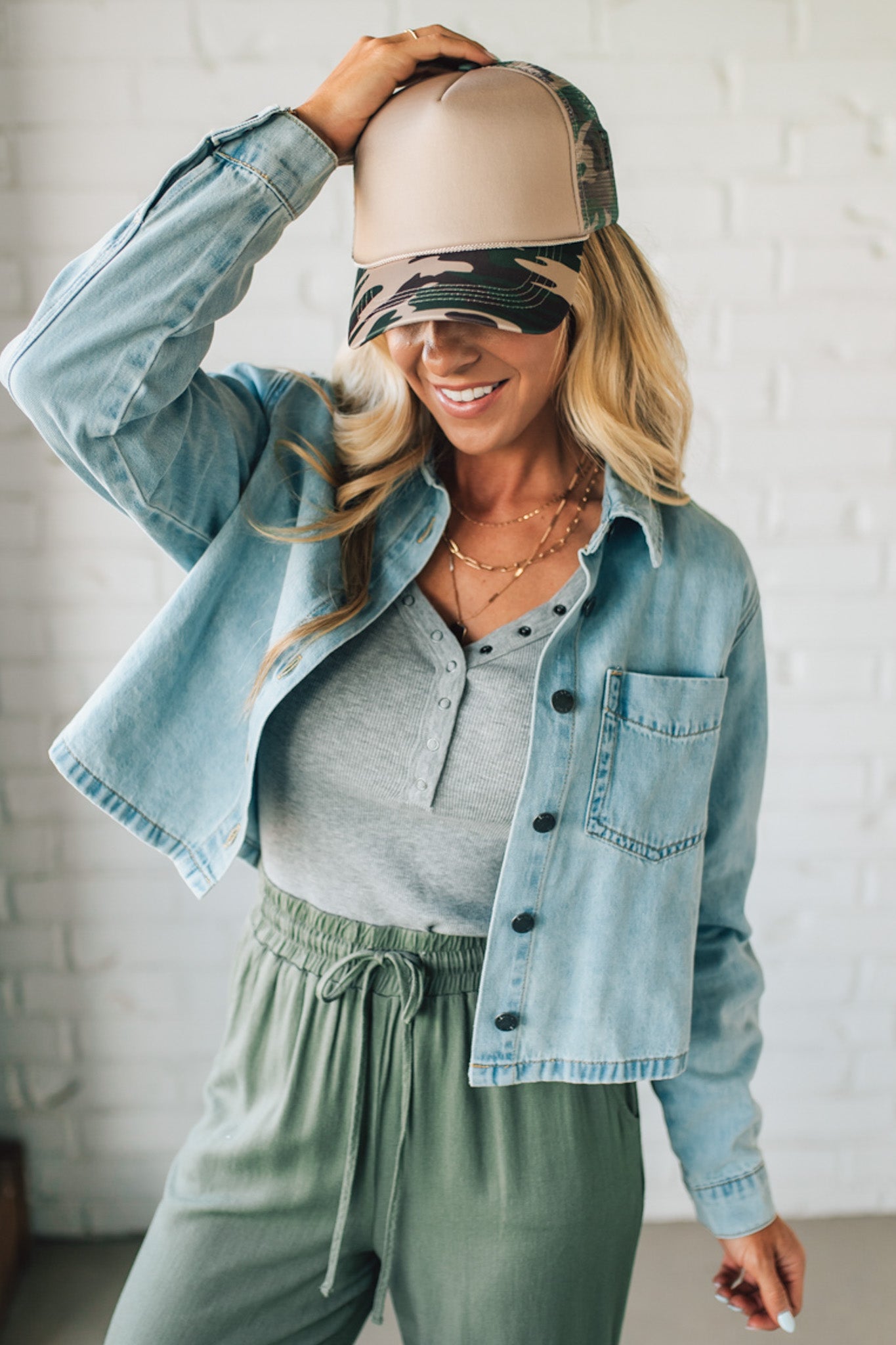 Woman wearing a denim jacket, gray top, green pants, and beige cap with visor.