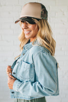 Woman wearing a beige and black cap and light blue denim jacket against a white brick wall.