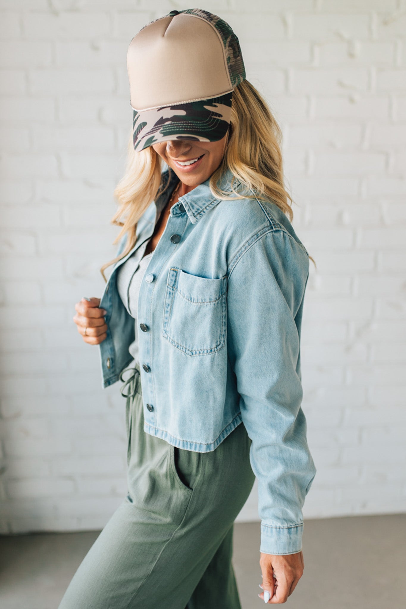 Woman wearing a denim jacket, green pants, and a camouflage visor against a white brick wall.