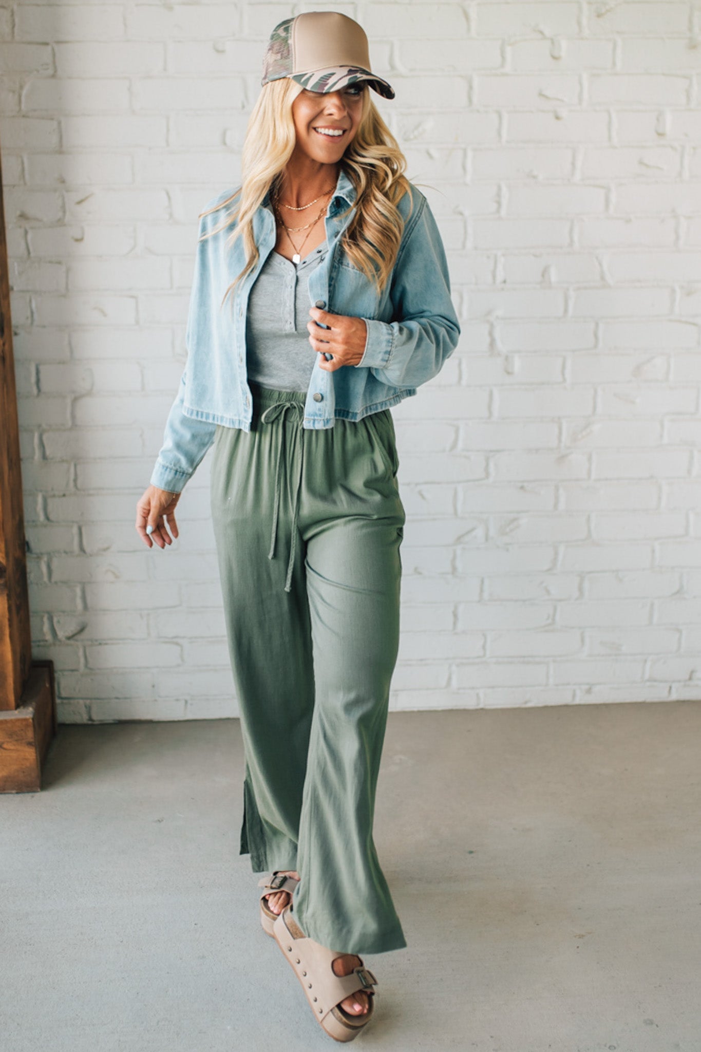 Woman wearing a light blue denim jacket, gray shirt, and green pants against a white brick wall.