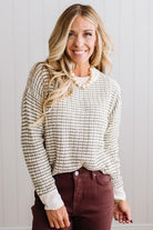 BLONDE WOMAN IN A BRIGHTLY LIT STUDIO WEARING A PAIR OF BROWN JEANS, CREAM CHUNKY BEADED NECKLACE AND A WAFFLED GRID SWEATER IN CREAM AND OLIVE GREEN.