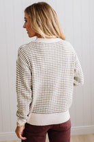 BLONDE WOMAN IN A BRIGHTLY LIT STUDIO WEARING A PAIR OF BROWN JEANS, CREAM CHUNKY BEADED NECKLACE AND A WAFFLED GRID SWEATER IN CREAM AND OLIVE GREEN.