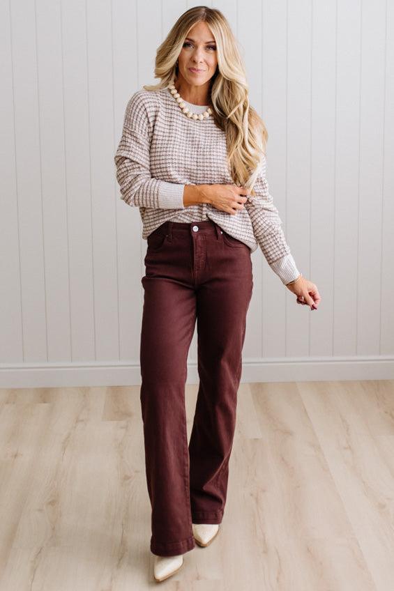 BLONDE WOMAN IN A BRIGHTLY LIT STUDIO WEARING A PAIR OF BROWN JEANS, CREAM CHUNKY BEADED NECKLACE AND A WAFFLED GRID SWEATER IN CREAM AND BROWN.