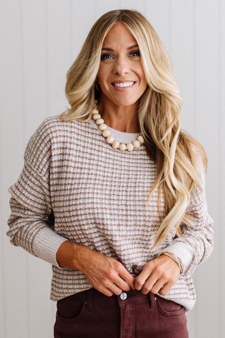 BLONDE WOMAN IN A BRIGHTLY LIT STUDIO WEARING A PAIR OF BROWN JEANS, CREAM CHUNKY BEADED NECKLACE AND A WAFFLED GRID SWEATER IN CREAM AND BROWN.