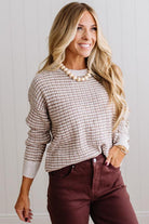 BLONDE WOMAN IN A BRIGHTLY LIT STUDIO WEARING A PAIR OF BROWN JEANS, CREAM CHUNKY BEADED NECKLACE AND A WAFFLED GRID SWEATER IN CREAM AND BROWN.
