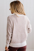BLONDE WOMAN IN A BRIGHTLY LIT STUDIO WEARING A PAIR OF BROWN JEANS, CREAM CHUNKY BEADED NECKLACE AND A WAFFLED GRID SWEATER IN CREAM AND BROWN.