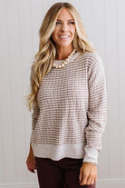 BLONDE WOMAN IN A BRIGHTLY LIT STUDIO WEARING A PAIR OF BROWN JEANS, CREAM CHUNKY BEADED NECKLACE AND A WAFFLED GRID SWEATER IN CREAM AND BROWN.