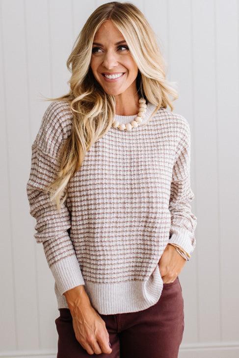 BLONDE WOMAN IN A BRIGHTLY LIT STUDIO WEARING A PAIR OF BROWN JEANS, CREAM CHUNKY BEADED NECKLACE AND A WAFFLED GRID SWEATER IN CREAM AND BROWN.