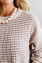 BLONDE WOMAN IN A BRIGHTLY LIT STUDIO WEARING A PAIR OF BROWN JEANS, CREAM CHUNKY BEADED NECKLACE AND A WAFFLED GRID SWEATER IN CREAM AND BROWN.