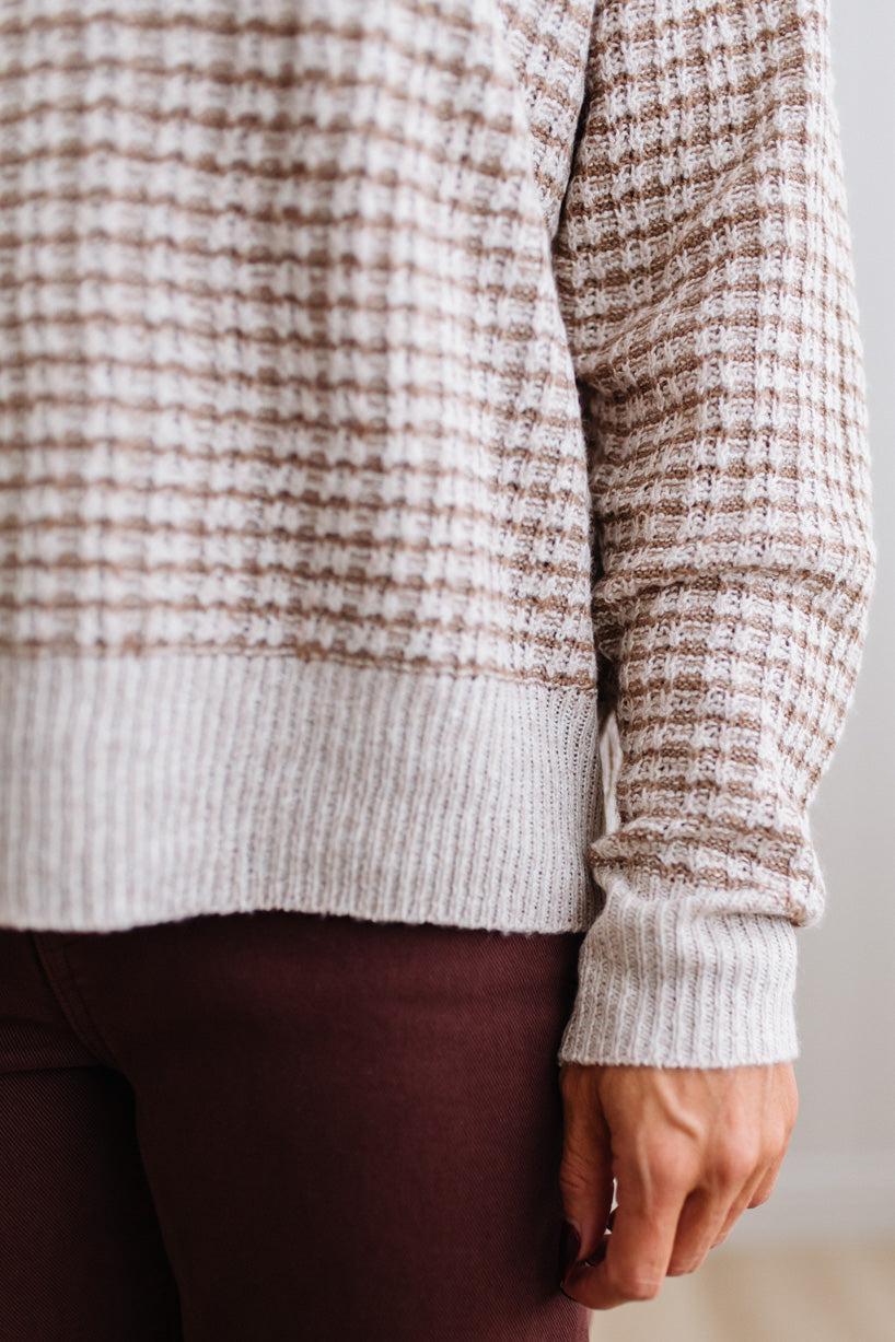 BLONDE WOMAN IN A BRIGHTLY LIT STUDIO WEARING A PAIR OF BROWN JEANS, CREAM CHUNKY BEADED NECKLACE AND A WAFFLED GRID SWEATER IN CREAM AND BROWN.