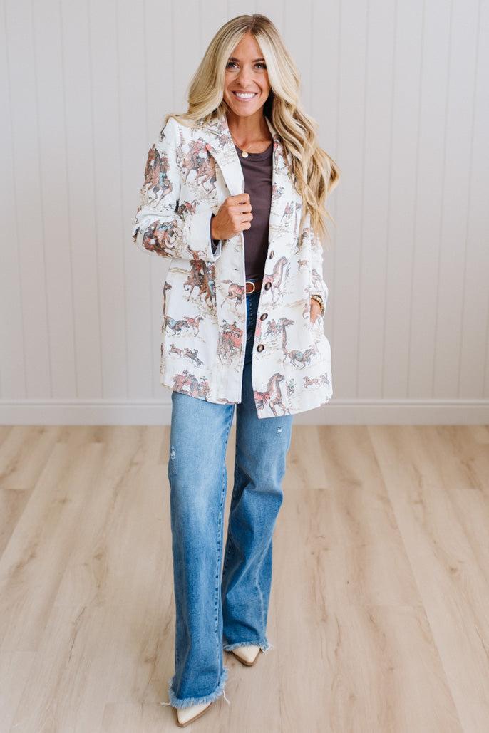 A woman modeling a white open-front blazer with a multicolor western-style horse print, paired with blue jeans and beige heels, standing in a room with light wood floors and white paneled walls.
