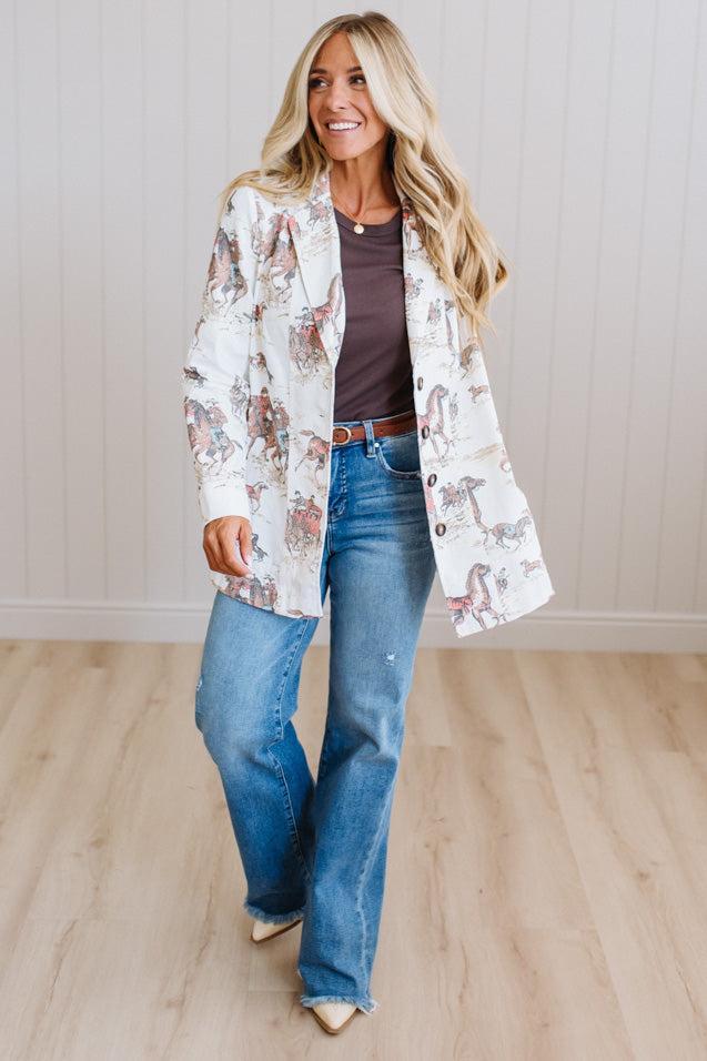 A woman modeling a long-sleeve western-style blazer with a white base and a multicolor horse print, worn over a dark brown top and blue jeans.