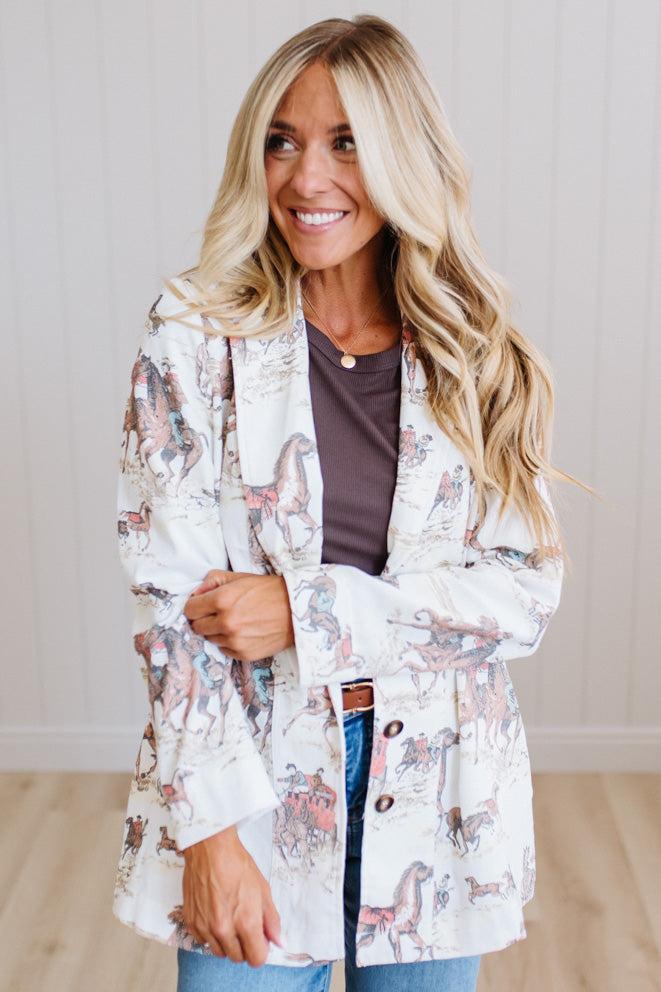 A woman with long blonde hair wearing a white button-up jacket with a pink and brown horse print, layered over a dark brown top and blue jeans, standing in front of a white paneled wall.