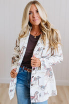 A woman modeling a white open-front blazer with a multicolor cowboy and cattle print, worn over a dark brown top and blue jeans.