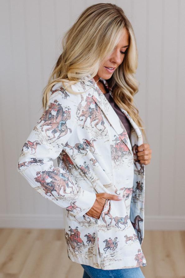 A woman wearing a white long-sleeve blazer with a detailed western-themed horse and rider print, paired with a dark top and blue jeans, standing in a brightly lit room with light wood floors and paneled walls.