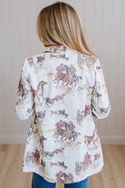 A woman viewed from behind wearing a white long-sleeve button-up shirt with a cowboy and horse print, paired with blue jeans, standing in front of a light wood-paneled wall.