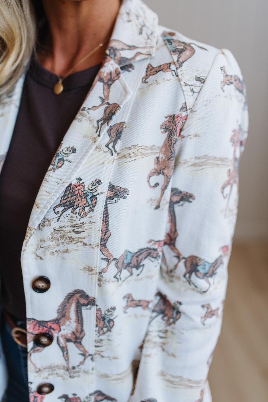 A close-up of a person wearing a white button-front blazer with an allover printed scene of horses and riders in muted brown, pink, and blue tones, layered over a dark brown top.