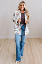 A woman modeling a white open-front blazer with a brown and beige floral print, paired with a dark brown top, blue jeans, and beige heels, standing in a room with light wood floors and white paneled walls.