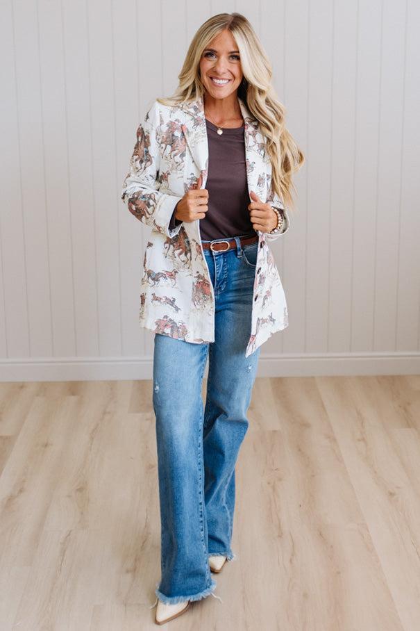 A woman modeling a white open-front blazer with a brown and beige floral print, paired with a dark brown top, blue jeans, and beige heels, standing in a room with light wood floors and white paneled walls.