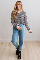 Blonde woman wearing light jeans and black boots with a black and white striped loose sweater with rust red contrast stitched trim. Standing on a light wood floor with paneled wall behind her.