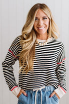 Blonde woman wearing light jeans and black boots with a black and white striped loose sweater with rust red contrast stitched trim. Standing on a light wood floor with paneled wall behind her.