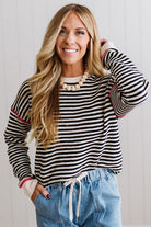 Blonde woman wearing light jeans and black boots with a black and white striped loose sweater with rust red contrast stitched trim. Standing on a light wood floor with paneled wall behind her.