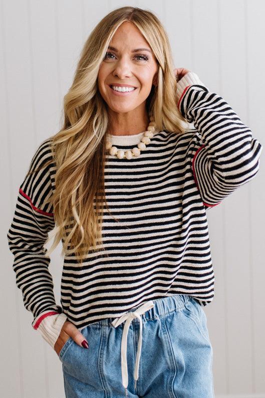 Blonde woman wearing light jeans and black boots with a black and white striped loose sweater with rust red contrast stitched trim. Standing on a light wood floor with paneled wall behind her.