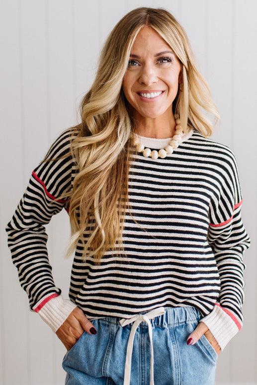 Blonde woman wearing light jeans and black boots with a black and white striped loose sweater with rust red contrast stitched trim. Standing on a light wood floor with paneled wall behind her.