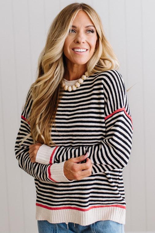 Blonde woman wearing light jeans and black boots with a black and white striped loose sweater with rust red contrast stitched trim. Standing on a light wood floor with paneled wall behind her.