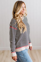Blonde woman wearing light jeans and black boots with a black and white striped loose sweater with rust red contrast stitched trim. Standing on a light wood floor with paneled wall behind her.