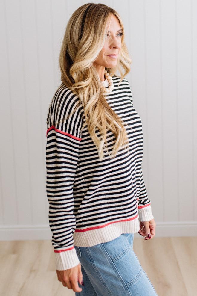 Blonde woman wearing light jeans and black boots with a black and white striped loose sweater with rust red contrast stitched trim. Standing on a light wood floor with paneled wall behind her.