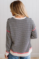 Blonde woman wearing light jeans and black boots with a black and white striped loose sweater with rust red contrast stitched trim. Standing on a light wood floor with paneled wall behind her.