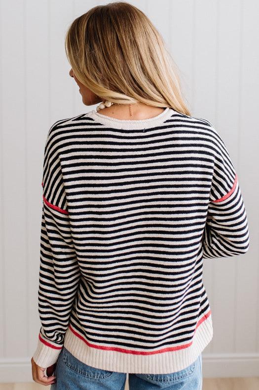 Blonde woman wearing light jeans and black boots with a black and white striped loose sweater with rust red contrast stitched trim. Standing on a light wood floor with paneled wall behind her.