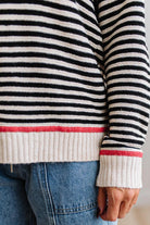 Blonde woman wearing light jeans and black boots with a black and white striped loose sweater with rust red contrast stitched trim. Standing on a light wood floor with paneled wall behind her.