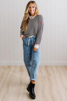 Blonde woman wearing light jeans and black boots with a black and white striped loose sweater with rust red contrast stitched trim. Standing on a light wood floor with paneled wall behind her.