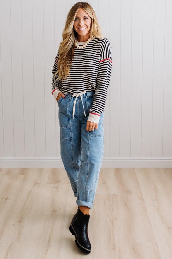 Blonde woman wearing light jeans and black boots with a black and white striped loose sweater with rust red contrast stitched trim. Standing on a light wood floor with paneled wall behind her.