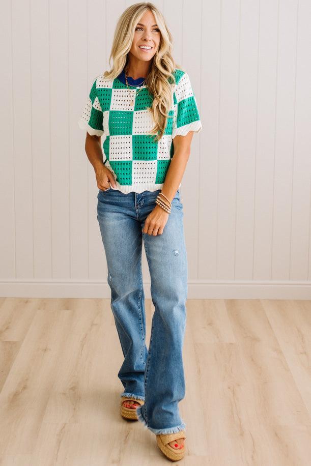 A woman modeling a green and white crochet checkered short-sleeve top with blue jeans and tan slide sandals, standing in a room with light wood floors and white paneled walls.