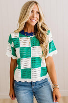 A woman wearing a green and white crochet checkered short-sleeve top with blue jeans, standing against a light-colored paneled wall.