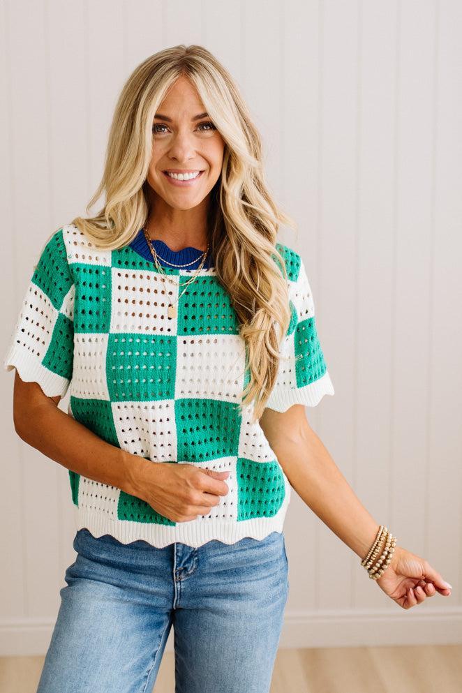 A woman wearing a green and white crochet checkerboard short-sleeve top with blue jeans, standing against a white paneled wall.