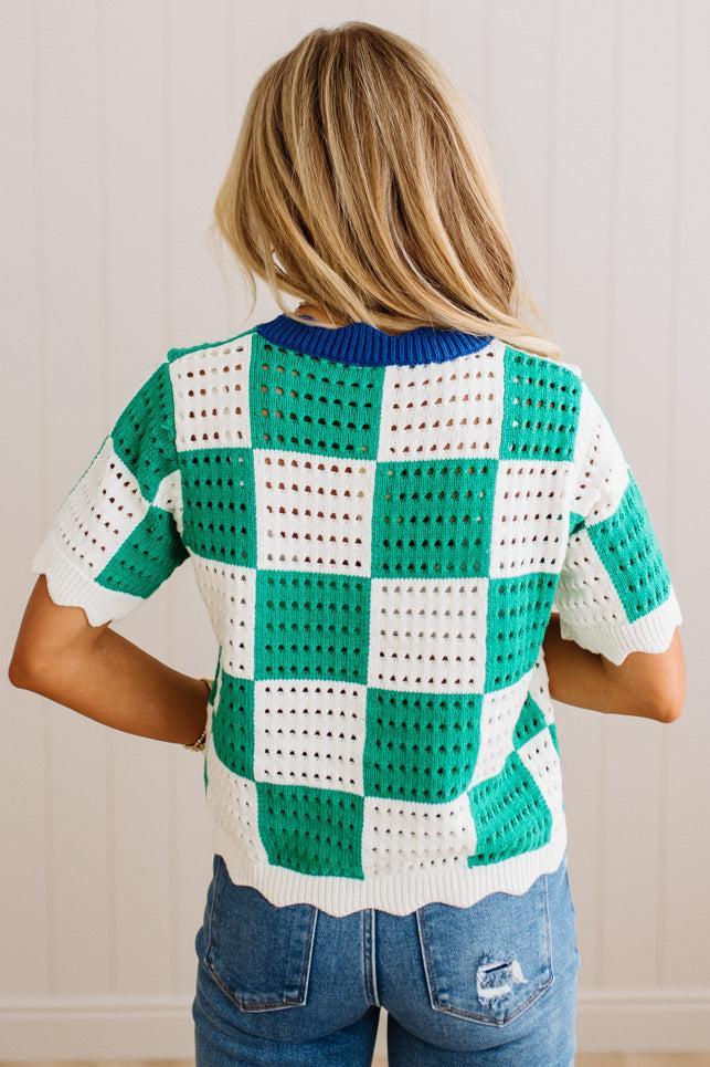 A woman viewed from the back wearing a short-sleeve knit top with a green and white checkerboard pattern and a blue ribbed collar, paired with blue jeans.