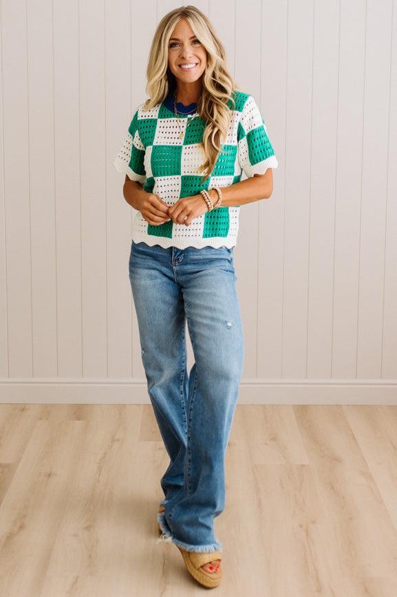 A woman modeling a green and white checkered crochet short-sleeve top with blue jeans and tan wedge sandals, standing against a white paneled wall and light wood floor.